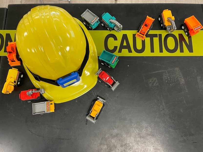 A construction hat on a table with caution tape, surrounded by toy trucks.