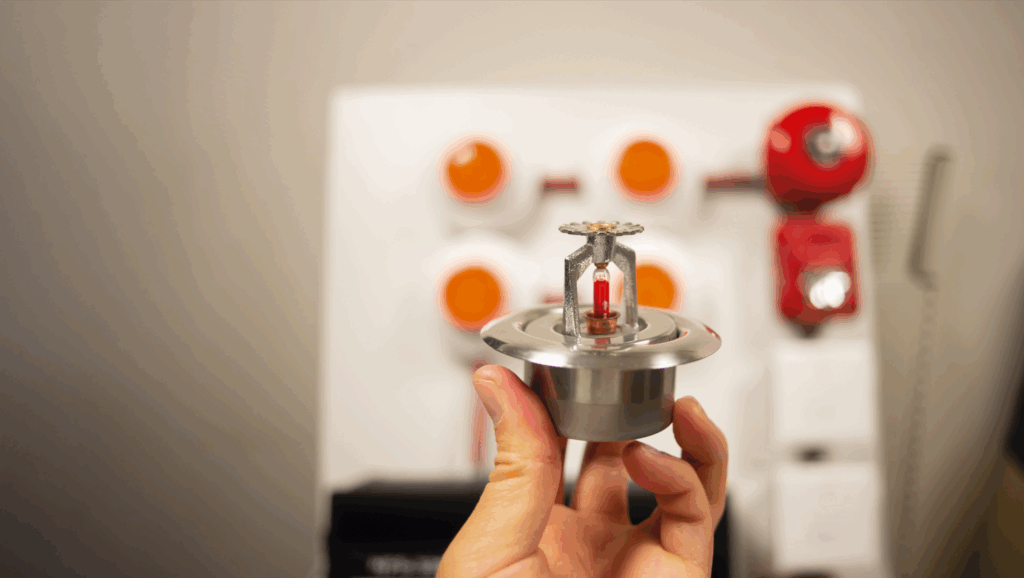 a hand holds a fire sprinkler with a classroom in the background
