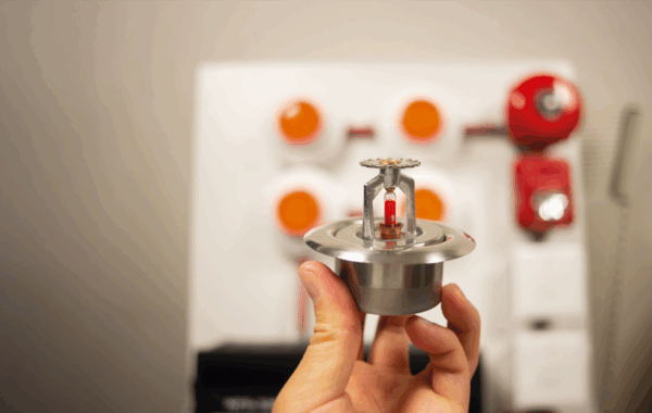 a hand holds a fire sprinkler with a classroom in the background
