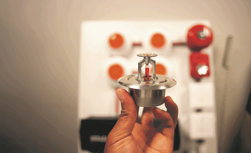 a hand holds a fire sprinkler with a classroom in the background