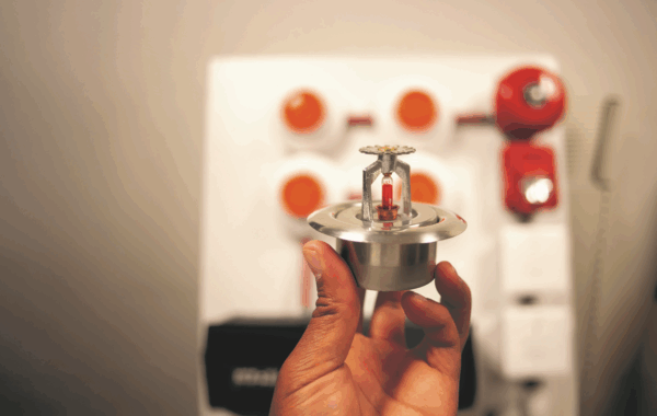 a hand holds a fire sprinkler with a classroom in the background