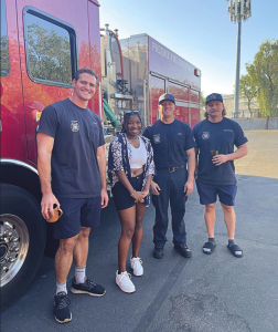 three fire professionals stand next to a fire truck, with student Jaleah Hamilton posing between them, smiling