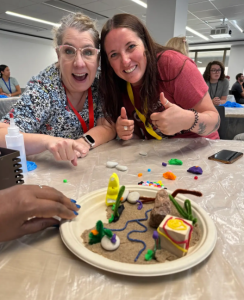 Nancy Schunke and a colleague pose with their thumbs up and smiling in front of an Xplorlabs cookie mining experiment, which shows cookie pieces on a plate on the table in front of them.