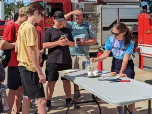Megan O'Keeffe wears an Xplorlabs t-shirt and leans over a table where she conducts a fire forensics investigation, while two middle school boys look on. A Cherokee County fire truck is pictured in the background.